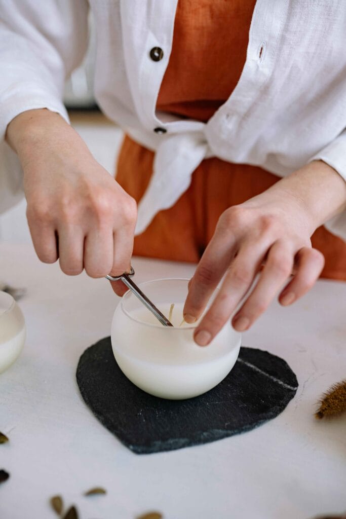 Person trimming a candle wick with scissors in a frosted glass jar, an essential step in how to make a candle wick for an even burn